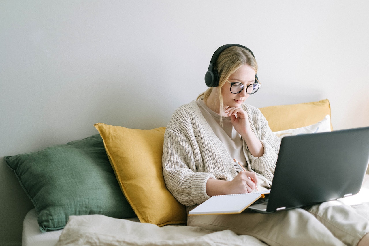 Young woman sitting on a bed with a laptop on her lap while writing in a notebook