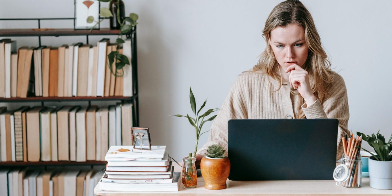A blonde woman working on her laptop at her desk with a full bookcase in the background