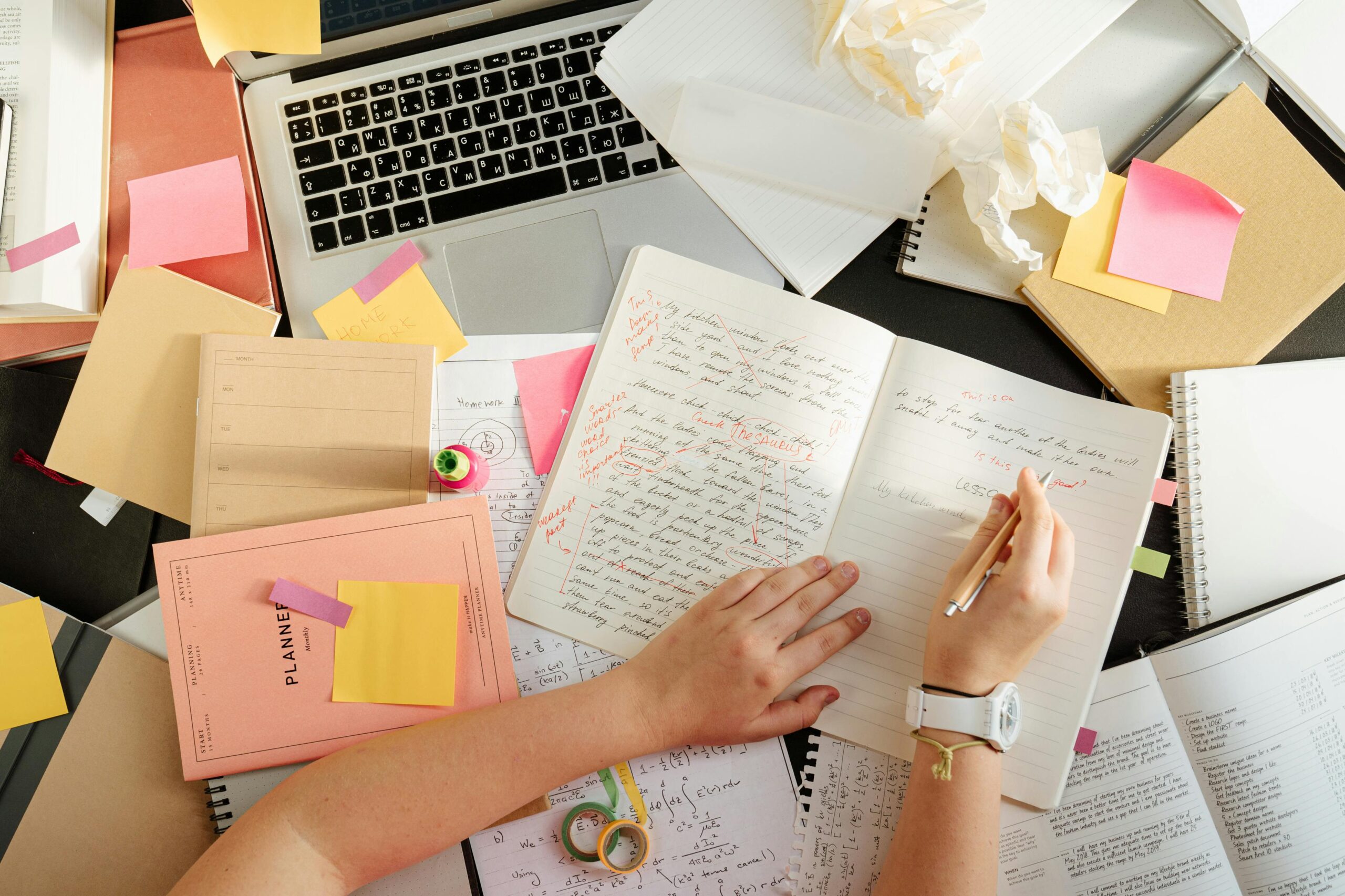 A messy desk covered in a laptop, papers, notebooks, and stationary.