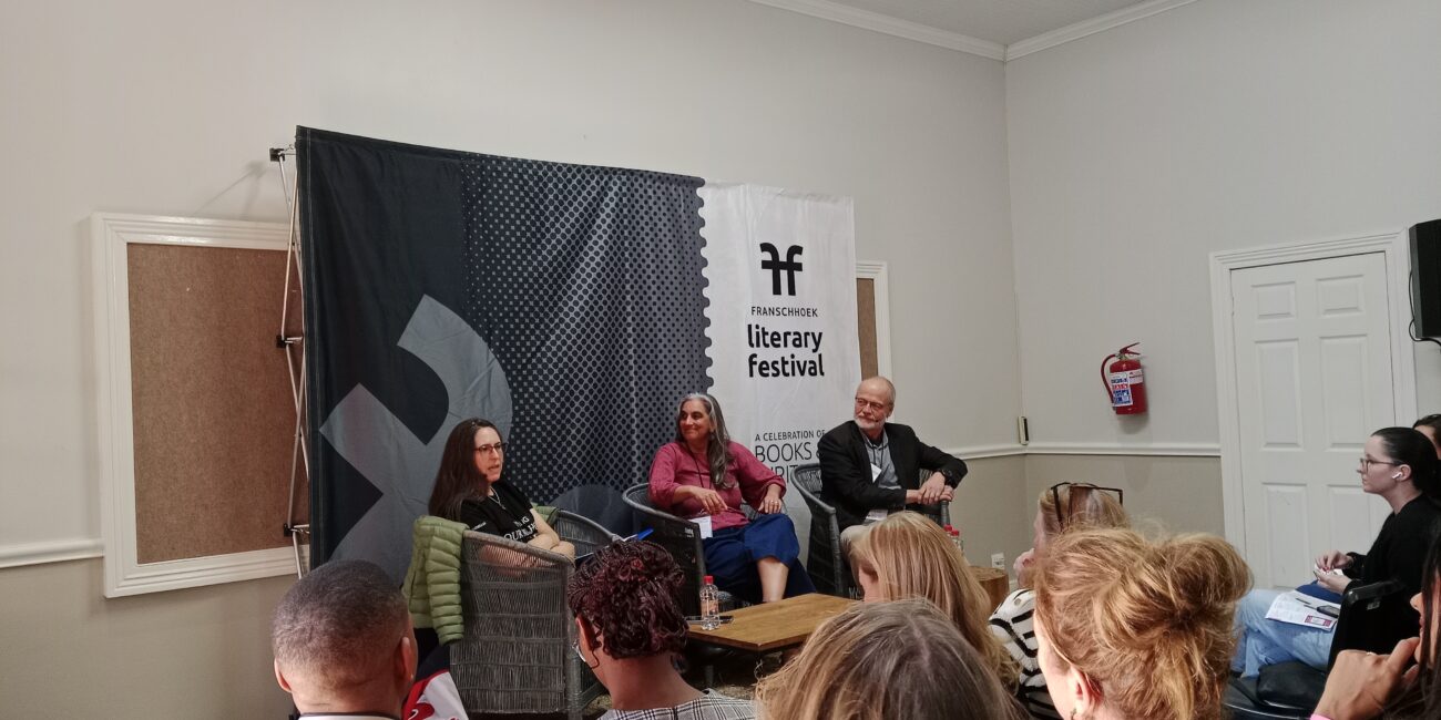 Two women and a man sitting on chairs on a stage. Behind them is a banner that reads Franschhoek Literary Festival.