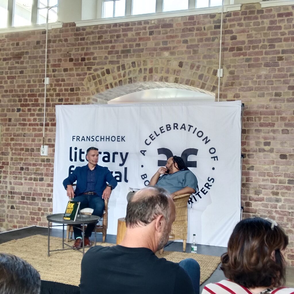 Two men sitting on chairs on a small stage. Behind them is a banner that reads Franschhoek Literary Festival.