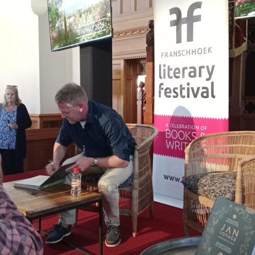 A man sitting in a chair in front of a table. The man is signing a book with a pen.